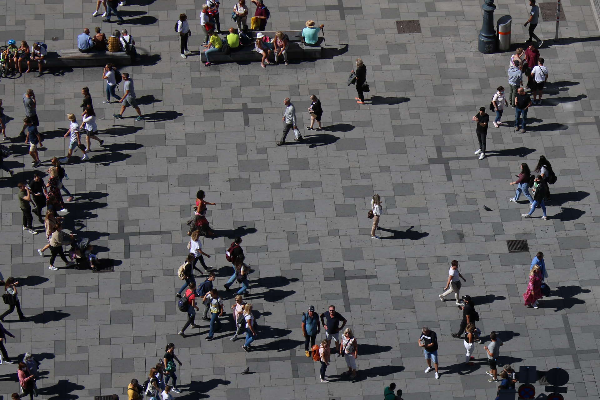 Vista cenital de una plaza urbana con numerosas personas y sus sombras alargadas.