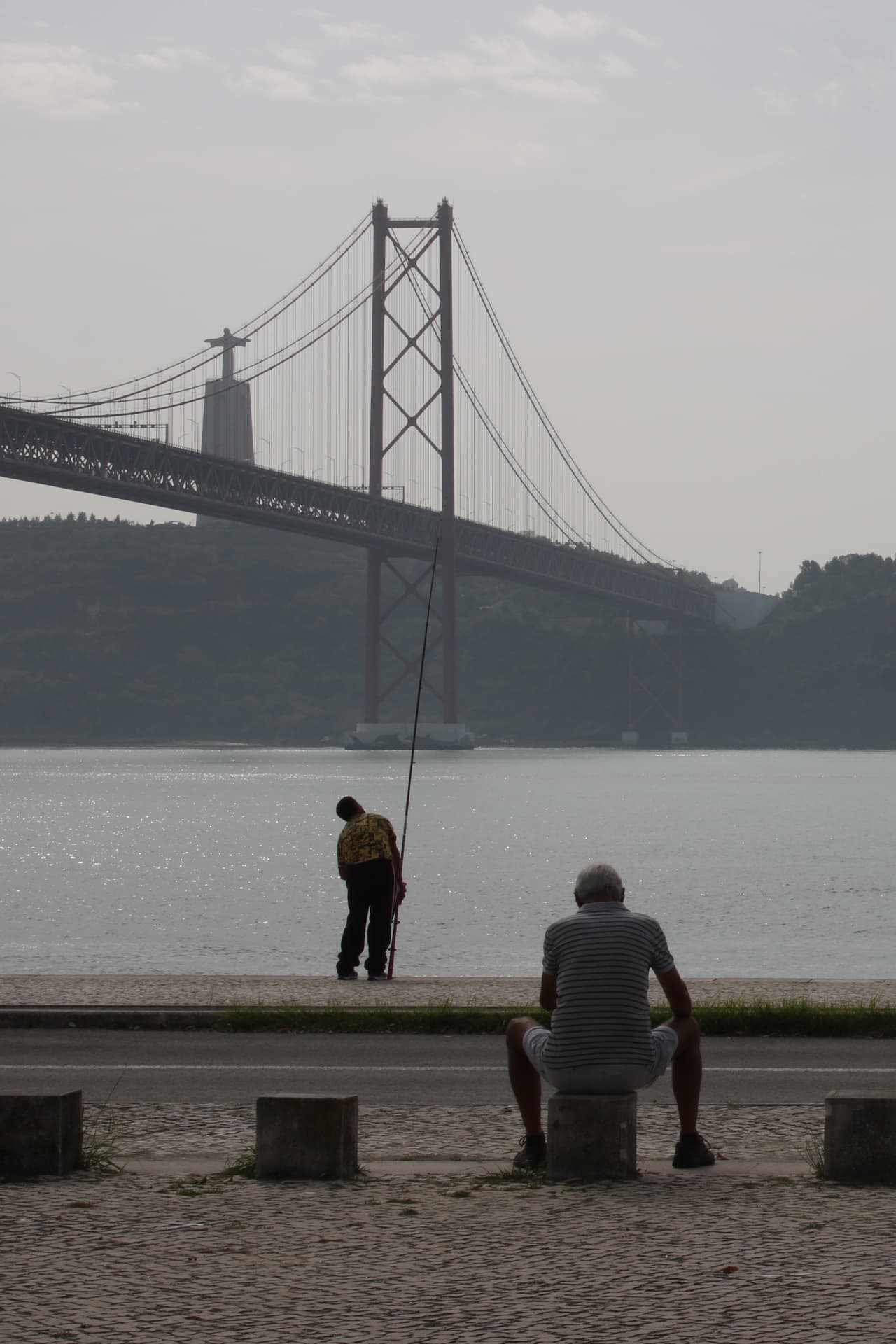 Dos figuras detenidas junto al agua con una gran infraestructura urbana al fondo en Lisboa.